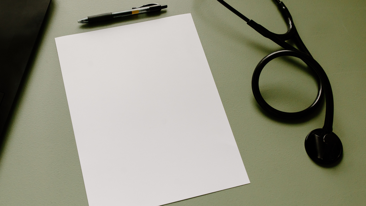 Photorealistic overhead view of a blank white sheet of paper beside a black pen and a stethoscope on a sage-green clinic desk in soft natural light