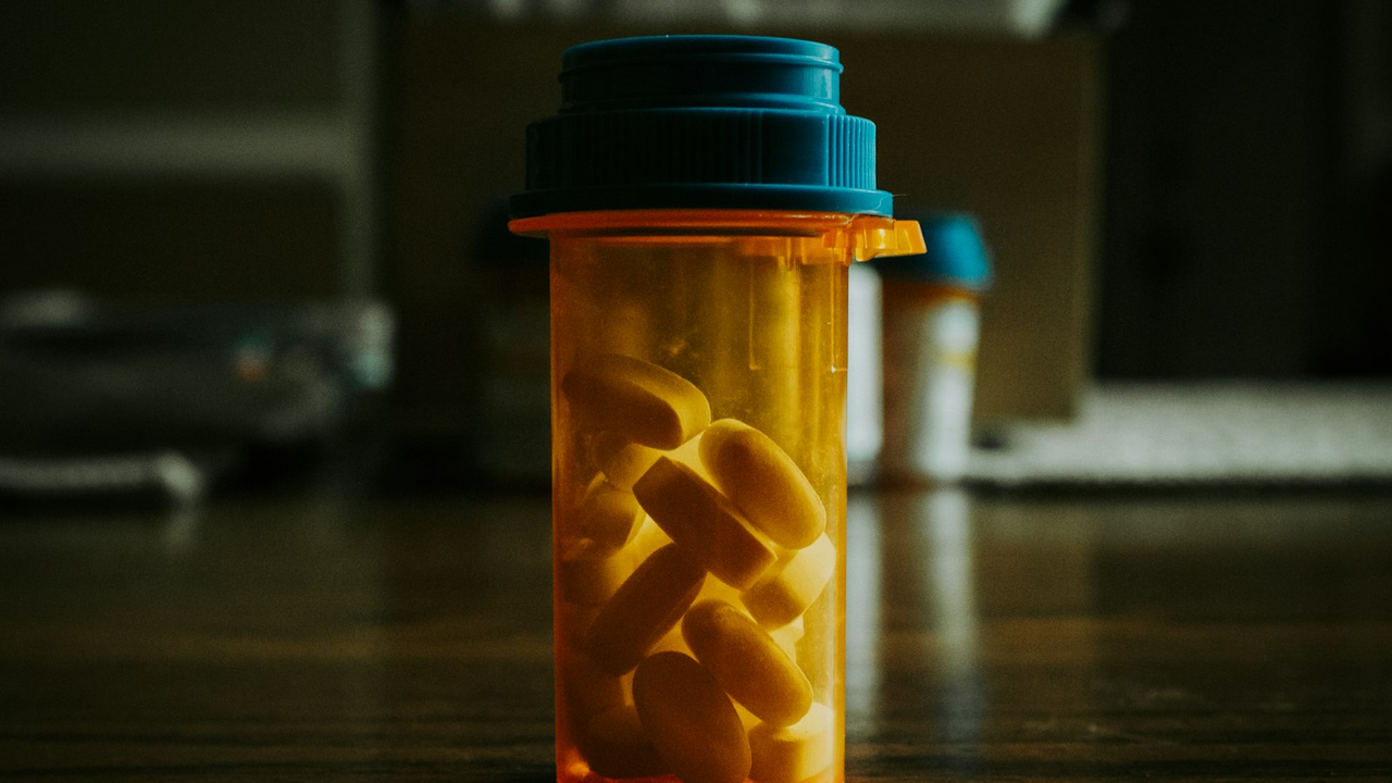 Photorealistic close-up of an unmarked amber medicine bottle with a teal child-resistant cap filled with white oblong tablets on a dark surface under soft side light, soft-focus bottles in the background